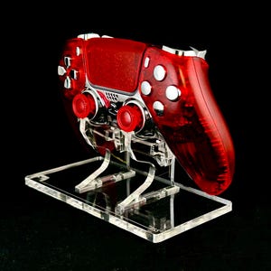 Puede incluir: Un mando de videojuegos rojo transparente descansa sobre un soporte de acrílico transparente. El mando tiene joysticks y botones rojos, con detalles plateados. El soporte es rectangular y transparente, para accesorios de juego.