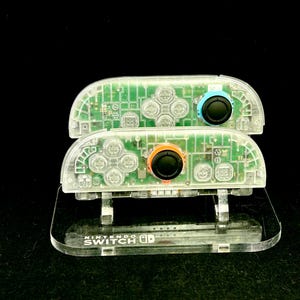 Könnte beinhalten: Zwei transparente Nintendo Switch Joy-Con-Controller, die interne Schaltkreise freilegen, auf einem transparenten Ständer gestapelt. Die Controller haben blaue und orangefarbene Knöpfe und schwarze Joysticks. Der Ständer trägt das Nintendo Switch-Logo.