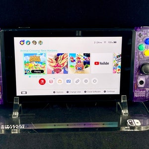Pu&ograve; includere: Una console Nintendo Switch viola con una custodia trasparente, che mostra il gioco Animal Crossing: New Horizons sullo schermo. La console &egrave; su un supporto in acrilico trasparente con il logo Nintendo Switch.