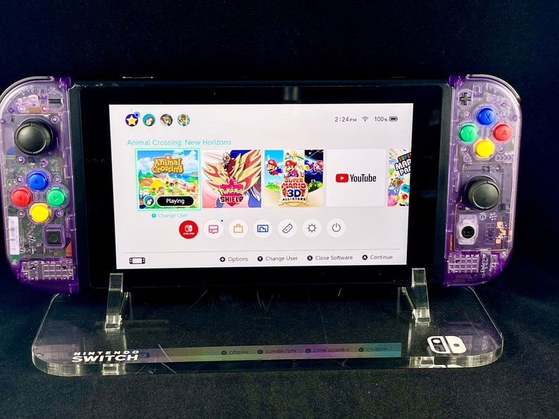 Pu&ograve; includere: Una console Nintendo Switch viola con una custodia trasparente, che mostra il gioco Animal Crossing: New Horizons sullo schermo. La console &egrave; su un supporto in acrilico trasparente con il logo Nintendo Switch.