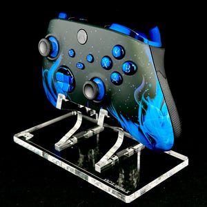 May include: A custom Xbox Series X controller with a black and blue design, displayed on a clear acrylic stand. The controller features blue buttons and accents, with a flame-like pattern. The stand is labeled "XBOX series X".