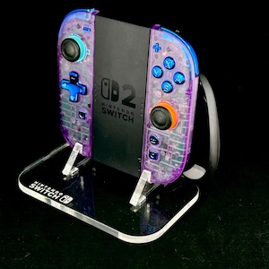 May include: A clear acrylic stand holding a Nintendo Switch console and controllers. The controllers have a transparent purple and blue design with colorful buttons. The stand has the Nintendo Switch logo.