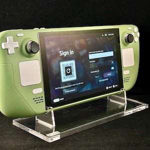 May include: A light green handheld gaming console on a clear acrylic stand. The screen displays a sign-in page with a QR code. The console has a D-pad, buttons, and joysticks. The screen prompts to create an account.