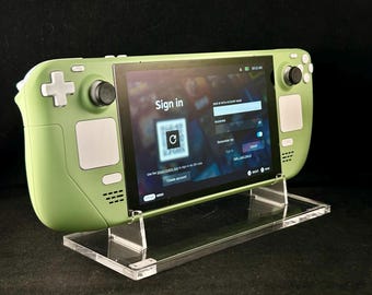 May include: A light green handheld gaming console with a screen displaying a sign-in page. The console is on a clear acrylic stand. The screen shows a QR code and prompts to create an account.
