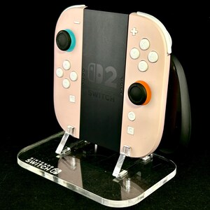 May include: A pink Nintendo Switch controller with white buttons and black joysticks, resting on a clear acrylic stand. The stand has the Nintendo Switch logo and supports the controller's black center section, which displays the Switch logo.