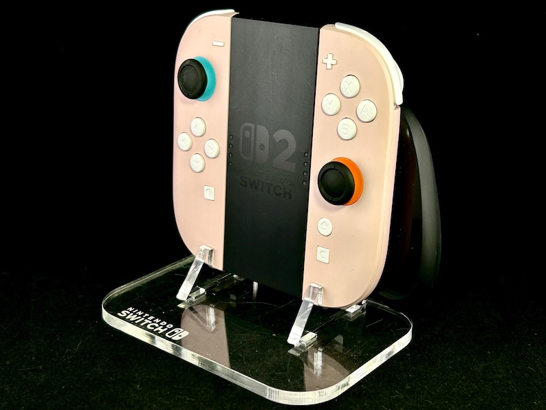 May include: A pink Nintendo Switch controller with white buttons and black joysticks, resting on a clear acrylic stand. The stand has the Nintendo Switch logo and supports the controller's black center section, which displays the Switch logo.