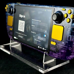 May include: A purple and gold Steam Deck handheld gaming console with a clear acrylic stand. The console is displaying a sign-in screen with a QR code and text that says "Sign in".