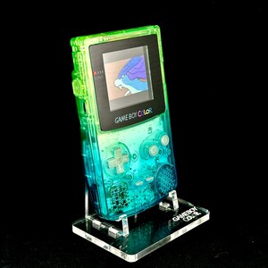 May include: A transparent green and blue Game Boy Color handheld console, displaying a game screen, is mounted on a clear acrylic stand. The stand has the words "Game Boy Color" etched on it. The background is black.