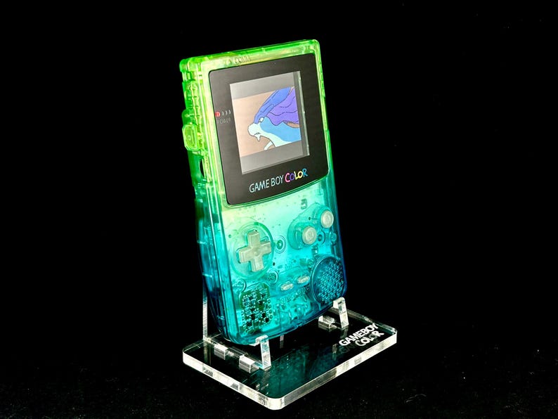 May include: A transparent green and blue Game Boy Color handheld console, displaying a game screen, is mounted on a clear acrylic stand. The stand has the words "Game Boy Color" etched on it. The background is black.