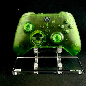 Puede incluir: Un mando Xbox Series X transparente verde en un soporte de acrílico transparente. El soporte tiene el texto "XBOX Series X" impreso en la parte delantera.