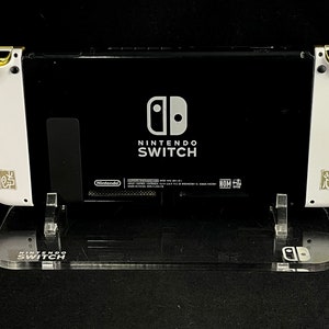 K&ouml;nnte beinhalten: Eine wei&szlig;e und schwarze Nintendo Switch-Konsole mit goldenen Akzenten an den Joy-Con-Controllern. Die Konsole steht auf einem durchsichtigen Acryl-St&auml;nder mit dem Nintendo Switch-Logo auf der Basis.