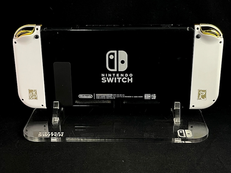 K&ouml;nnte beinhalten: Eine wei&szlig;e und schwarze Nintendo Switch-Konsole mit goldenen Akzenten an den Joy-Con-Controllern. Die Konsole steht auf einem durchsichtigen Acryl-St&auml;nder mit dem Nintendo Switch-Logo auf der Basis.