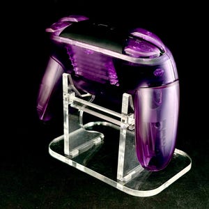 Op de afbeelding: Een transparante paarse videogamecontroller op een heldere acrylstandaard. De controller heeft een zwarte bovenkant en een doorzichtige behuizing. De standaard is ook helder en heeft een rechthoekige basis.
