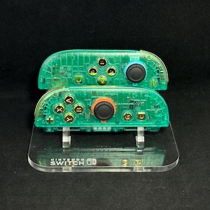 Pode incluir: Dois comandos Joy-Con Nintendo Switch verdes transparentes empilhados em um suporte de acrílico transparente. Os comandos têm botões dourados e manípulos analógicos pretos. O suporte tem o logotipo Nintendo Switch.
