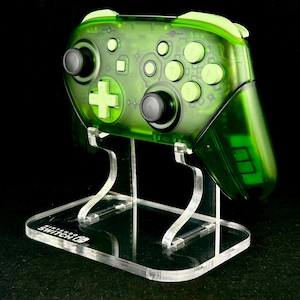 以下が含まれることがあります： 透明な緑色のNintendo Switch Proコントローラーが、透明なアクリルスタンドに置かれています。コントローラーには黒と緑のボタンと十字キーがあります。スタンドにはNintendo Switchのロゴが表示されています。