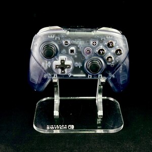 Puede incluir: Un mando transparente de Nintendo Switch con botones negros y una cruceta, apoyado en un soporte de acr&iacute;lico transparente. El soporte tiene el logotipo de Nintendo Switch. Los componentes internos del mando son visibles.