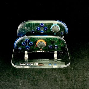 May include: Clear acrylic Nintendo Switch Joy-Con controllers with visible internal circuitry. The controllers have blue and orange accents, with blue buttons. The base features the Nintendo Switch logo.