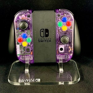 Pu&ograve; includere: Una console Nintendo Switch trasparente viola con un supporto in acrilico trasparente. La console ha una scocca trasparente viola con componenti interni visibili e pulsanti colorati. Il supporto ha la scritta "Nintendo Switch" stampata.
