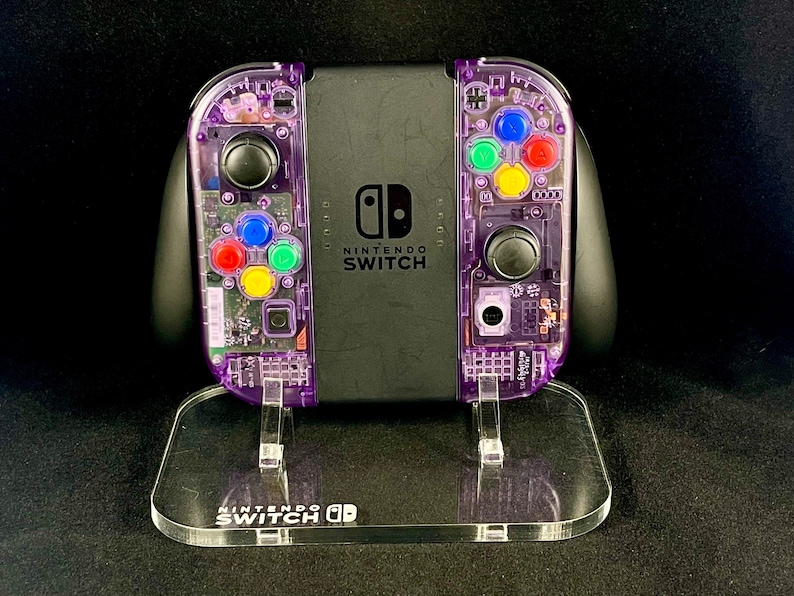 Pu&ograve; includere: Una console Nintendo Switch trasparente viola con un supporto in acrilico trasparente. La console ha una scocca trasparente viola con componenti interni visibili e pulsanti colorati. Il supporto ha la scritta "Nintendo Switch" stampata.