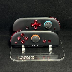 May include: Two black Nintendo Switch Joy-Con controllers with red accents and buttons, resting on a clear acrylic stand. The stand features the Nintendo Switch logo. One controller has a blue joystick, the other an orange one.
