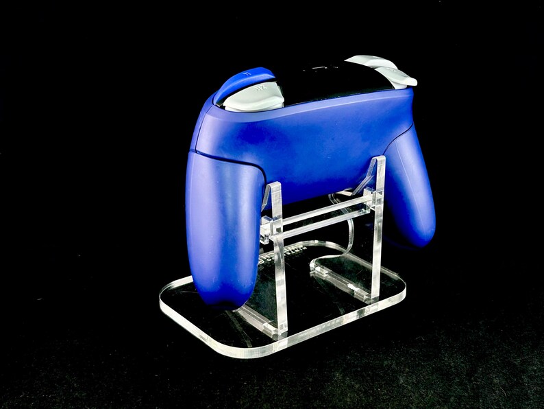 K&ouml;nnte beinhalten: Ein blauer Videospiel-Controller steht auf einem transparenten Acrylst&auml;nder. Der Controller hat wei&szlig;e und schwarze Akzente. Der St&auml;nder ist rechteckig mit abgerundeten Kanten und wird von transparenten, vertikalen St&uuml;tzen getragen.