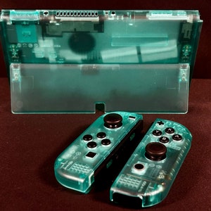 May include: A pair of transparent teal Nintendo Switch Joy-Con controllers with black buttons. The controllers are separated and lying on a dark brown surface.