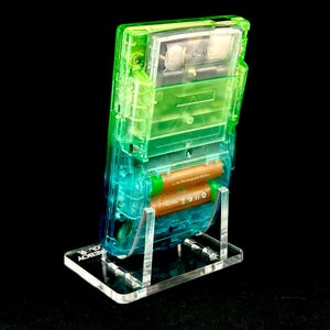 May include: A transparent green and blue Game Boy Color handheld video game console, with two brown batteries, is displayed on a clear acrylic stand. The stand has the words "GAMEBOY COLOR" etched on the base.