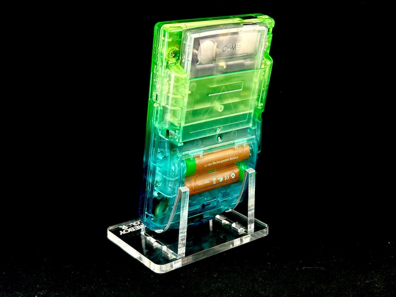 May include: A transparent green and blue Game Boy Color handheld video game console, with two brown batteries, is displayed on a clear acrylic stand. The stand has the words "GAMEBOY COLOR" etched on the base.
