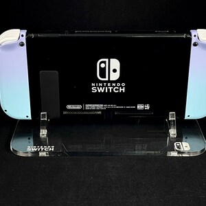 May include: A black Nintendo Switch console with a light blue and purple gradient on the sides. The console is on a clear acrylic stand with the words "Nintendo Switch" printed on the front.