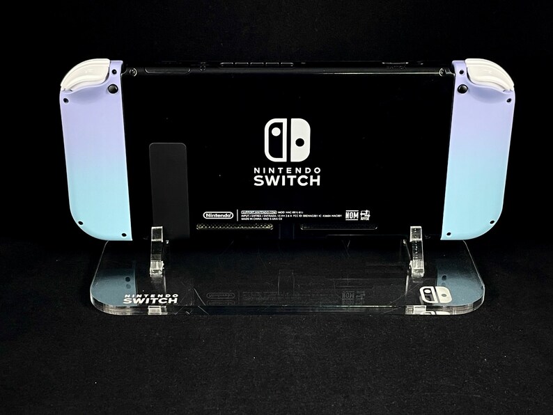May include: A black Nintendo Switch console with a light blue and purple gradient on the sides. The console is on a clear acrylic stand with the words "Nintendo Switch" printed on the front.