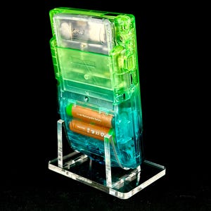 May include: A transparent green and blue Game Boy handheld console, showcasing its internal components, is displayed on a clear acrylic stand. The device contains two brown batteries. The background is black.