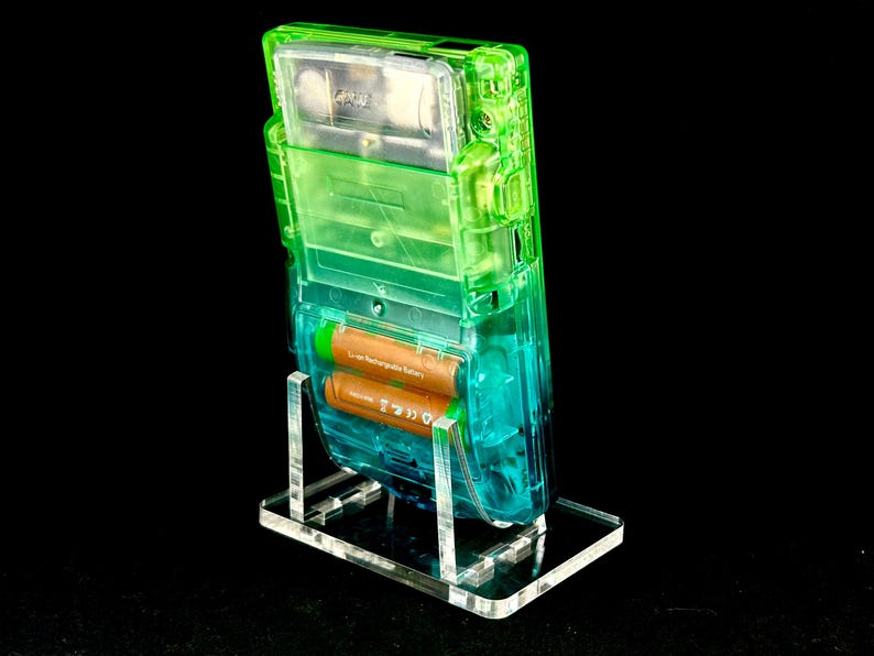 May include: A transparent green and blue Game Boy handheld console, showcasing its internal components, is displayed on a clear acrylic stand. The device contains two brown batteries. The background is black.