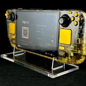 May include: A transparent yellow and black handheld gaming device with a screen displaying a sign-in prompt. The device is mounted on a clear acrylic stand. The buttons and controls are black and yellow.