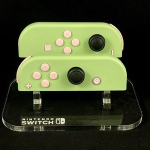 May include: Two light green Nintendo Switch Joy-Con controllers with pink buttons and black joysticks are displayed on a clear acrylic stand. The stand has the Nintendo Switch logo printed on it. The controllers are stacked on top of each other.