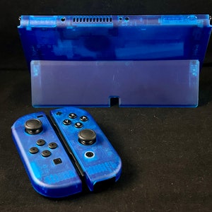 Könnte beinhalten: Eine durchsichtige blaue Nintendo Switch Konsole mit passenden blauen Joy-Con Controllern. Die Konsole ist geschlossen, wobei der Bildschirm zur Kamera zeigt. Die Joy-Cons sind abgenommen und liegen auf einer schwarzen Oberfläche.
