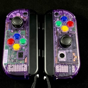 Pu&ograve; includere: Un paio di controller Nintendo Switch Joy-Con trasparenti viola con pulsanti neri e un joystick nero. I controller sono collegati tra loro e hanno un cinturino nero.