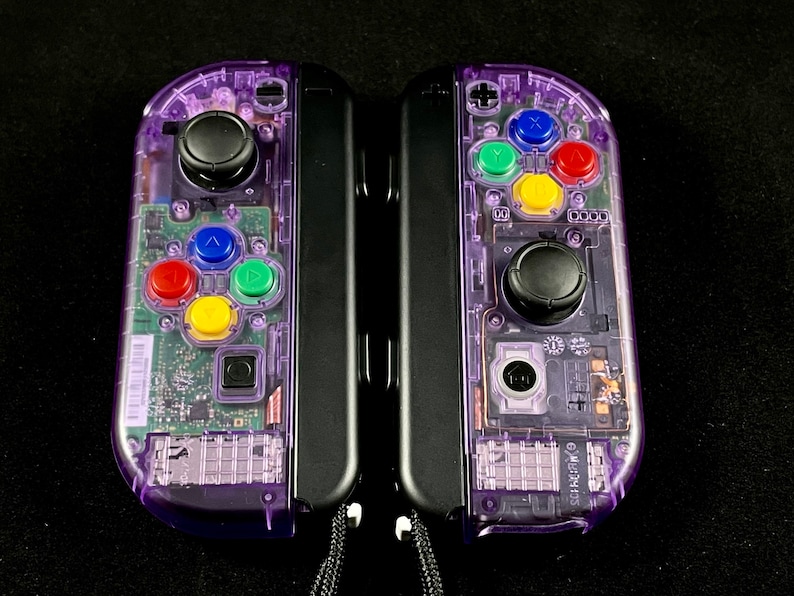 Pu&ograve; includere: Un paio di controller Nintendo Switch Joy-Con trasparenti viola con pulsanti neri e un joystick nero. I controller sono collegati tra loro e hanno un cinturino nero.