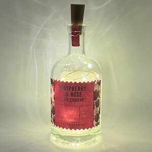 May include: A clear glass bottle with a cork stopper and a label that reads "Raspberry & Rose Gin Liqueur" with a pink and white floral design. The bottle is filled with white fairy lights.
