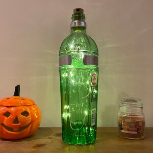 Pu&ograve; includere: Una bottiglia di vetro verde con un tappo di sughero e luci a stringa all'interno. La bottiglia &egrave; appoggiata su una superficie di legno accanto a una piccola zucca arancione e un barattolo di vetro con una candela all'interno.