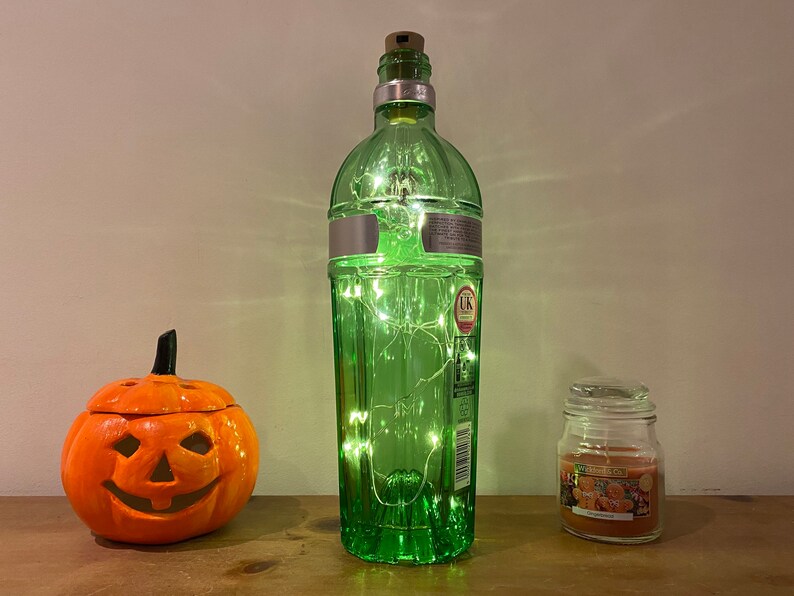 Pu&ograve; includere: Una bottiglia di vetro verde con un tappo di sughero e luci a stringa all'interno. La bottiglia &egrave; appoggiata su una superficie di legno accanto a una piccola zucca arancione e un barattolo di vetro con una candela all'interno.