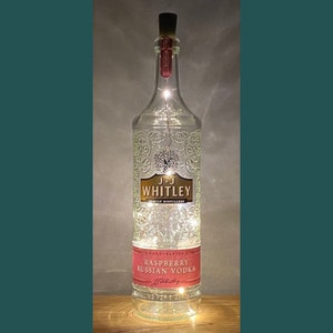 May include: A clear glass bottle with a cork top and a label that reads "Raspberry Russian Vodka" and "J.J. Whitley" with a decorative design. The bottle is lit with white fairy lights.