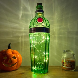 Pu&ograve; includere: Una bottiglia di vetro verde con un tappo di sughero e un'etichetta che recita "Tanqueray No Ten" &egrave; piena di luci bianche fatate. La bottiglia &egrave; appoggiata su una superficie di legno accanto a una piccola zucca di ceramica arancione e un barattolo di vetro con una candela.