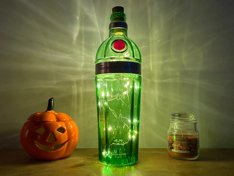 Pu&ograve; includere: Una bottiglia di vetro verde con un tappo di sughero e un'etichetta che recita "Tanqueray No Ten" &egrave; piena di luci bianche fatate. La bottiglia &egrave; appoggiata su una superficie di legno accanto a una piccola zucca di ceramica arancione e un barattolo di vetro con una candela.