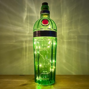 Pu&ograve; includere: Una bottiglia di vetro verde con un tappo di sughero e un'etichetta che recita "Tanqueray No. Ten" &egrave; piena di luci bianche fatate. La bottiglia &egrave; appoggiata su una superficie di legno.
