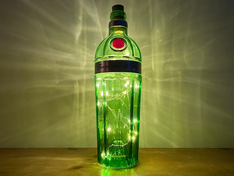 Pu&ograve; includere: Una bottiglia di vetro verde con un tappo di sughero e un'etichetta che recita "Tanqueray No. Ten" &egrave; piena di luci bianche fatate. La bottiglia &egrave; appoggiata su una superficie di legno.