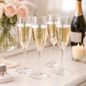 May include: Four champagne flutes filled with sparkling wine, with the words "Bride", "Bridesmaid", and "Maid of Honour" printed on them. A bottle of champagne and a lit candle are in the background.