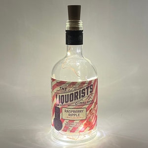 May include: A clear glass bottle with a cork stopper and a red, white, and black label. The label reads "The Liquourists Small Batch Raspberry Ripple Gin Liqueur".