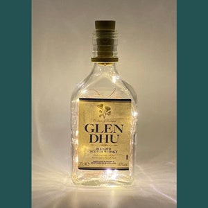 May include: A clear glass bottle with a cork stopper and a gold label that reads "Glen Dhu Blended Scotch Whisky". The bottle is decorated with white fairy lights.