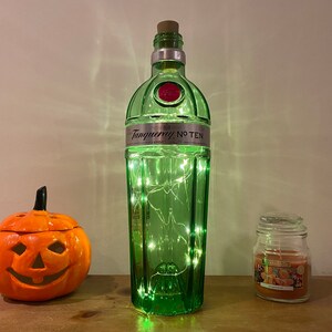Pu&ograve; includere: Una bottiglia di vetro verde con un tappo di sughero e luci bianche all'interno. La bottiglia ha l'etichetta "Tanqueray No Ten". C'&egrave; una piccola zucca arancione con una faccia di zucca e un barattolo di vetro con una candela dentro.