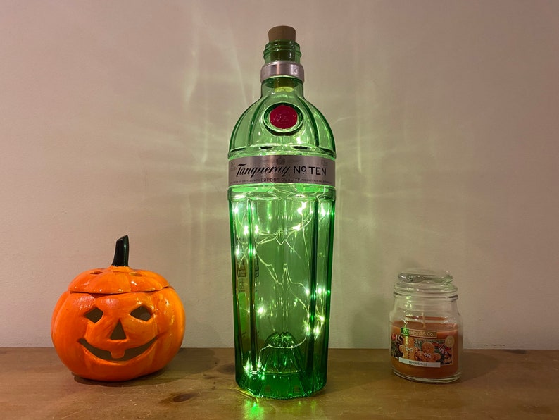Pu&ograve; includere: Una bottiglia di vetro verde con un tappo di sughero e luci bianche all'interno. La bottiglia ha l'etichetta "Tanqueray No Ten". C'&egrave; una piccola zucca arancione con una faccia di zucca e un barattolo di vetro con una candela dentro.